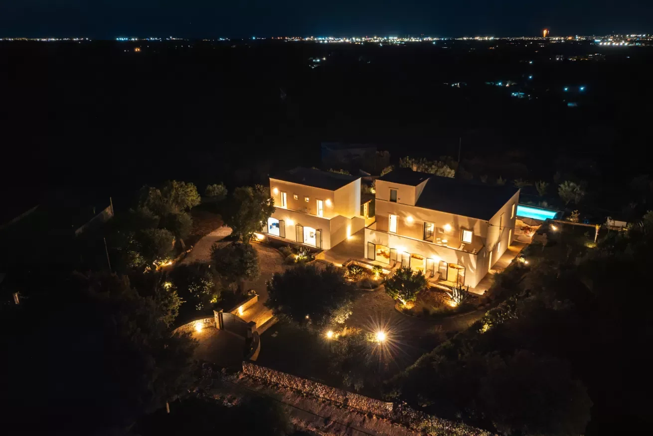 Aerial night view of a modern house with illuminated exterior lights and a swimming pool in the backyard, surrounded by trees and situated in a dark landscape with city lights in the distance.