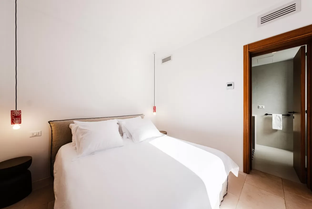 A modern bedroom with a white bed, white pillows, two red pendant lights, and an open bathroom door revealing a beige wall and a white towel on a black towel bar.