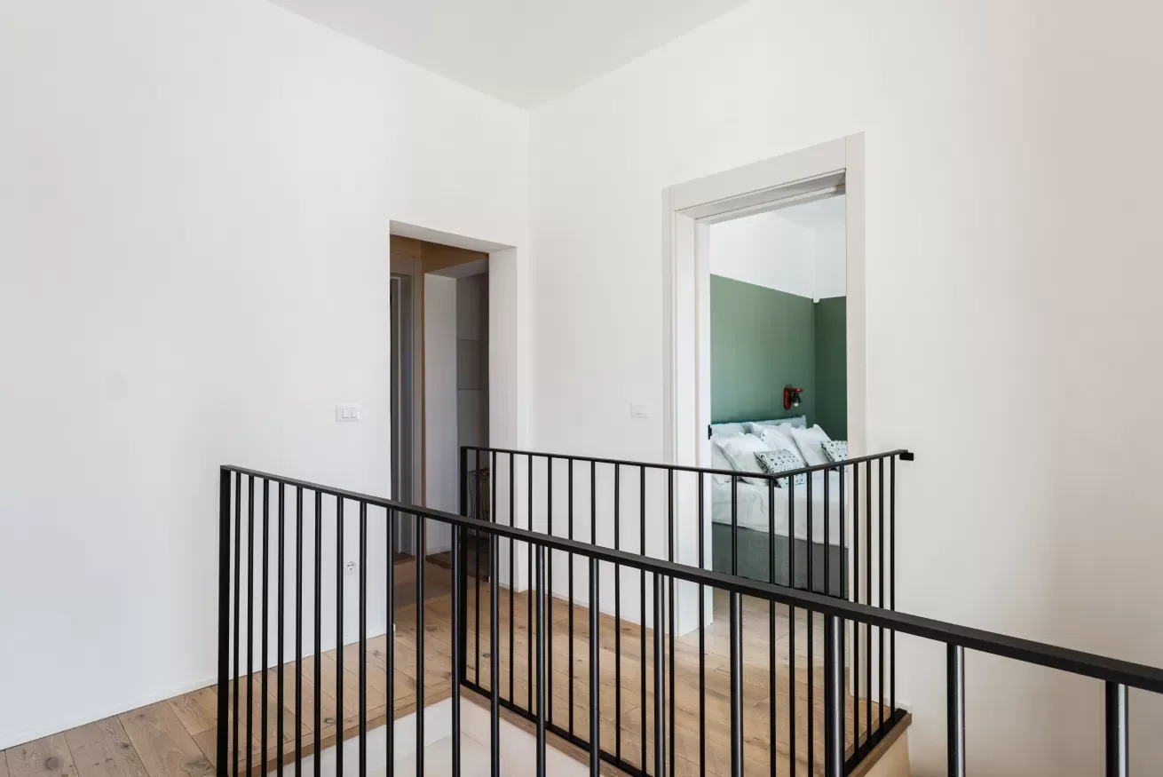 Interior view of a staircase landing with black metal railing, overlooking a hallway and a doorway leading to a bedroom with a green wall and white bedding.
