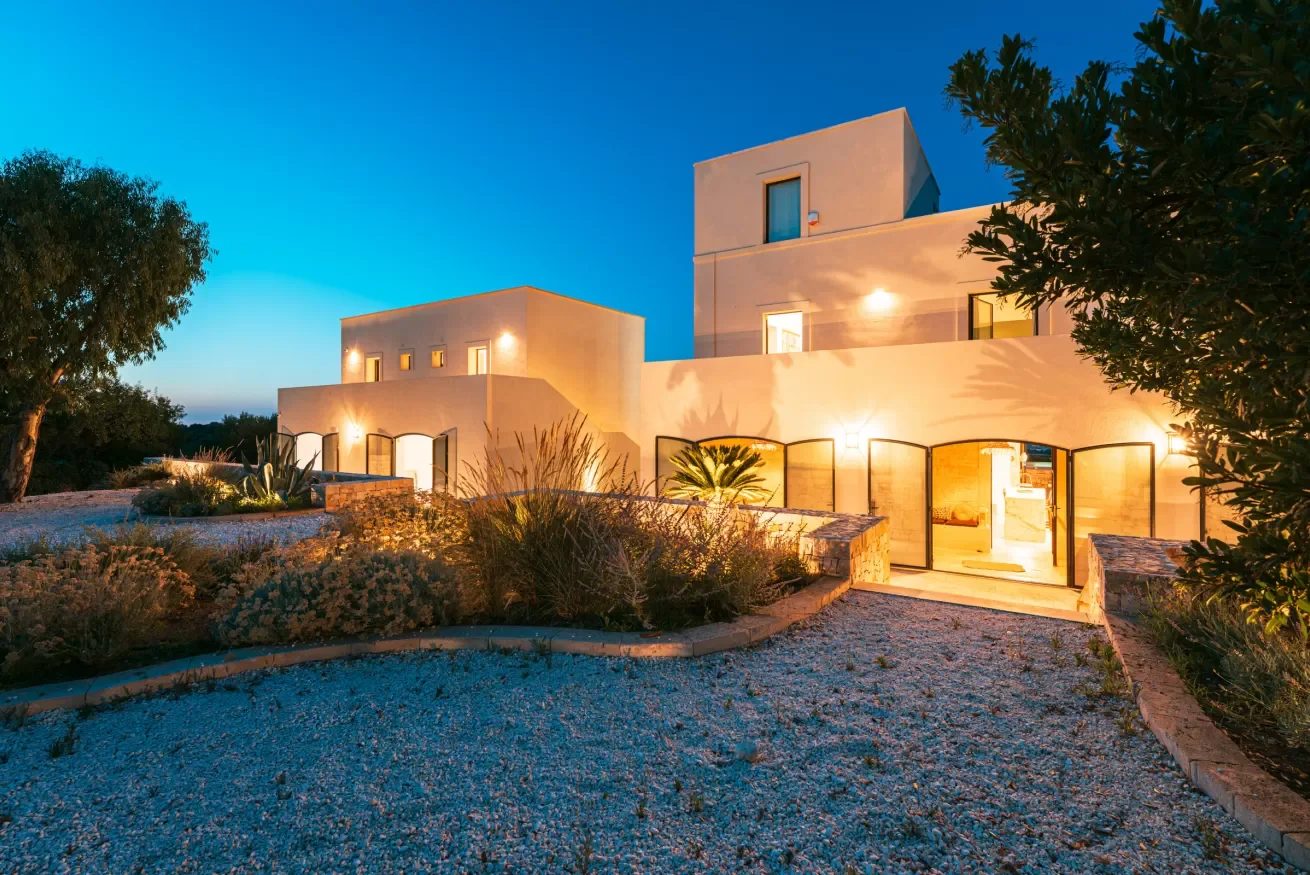 Modern white multi-story house with large glass doors and outdoor lighting, surrounded by desert landscaping, under a twilight sky.