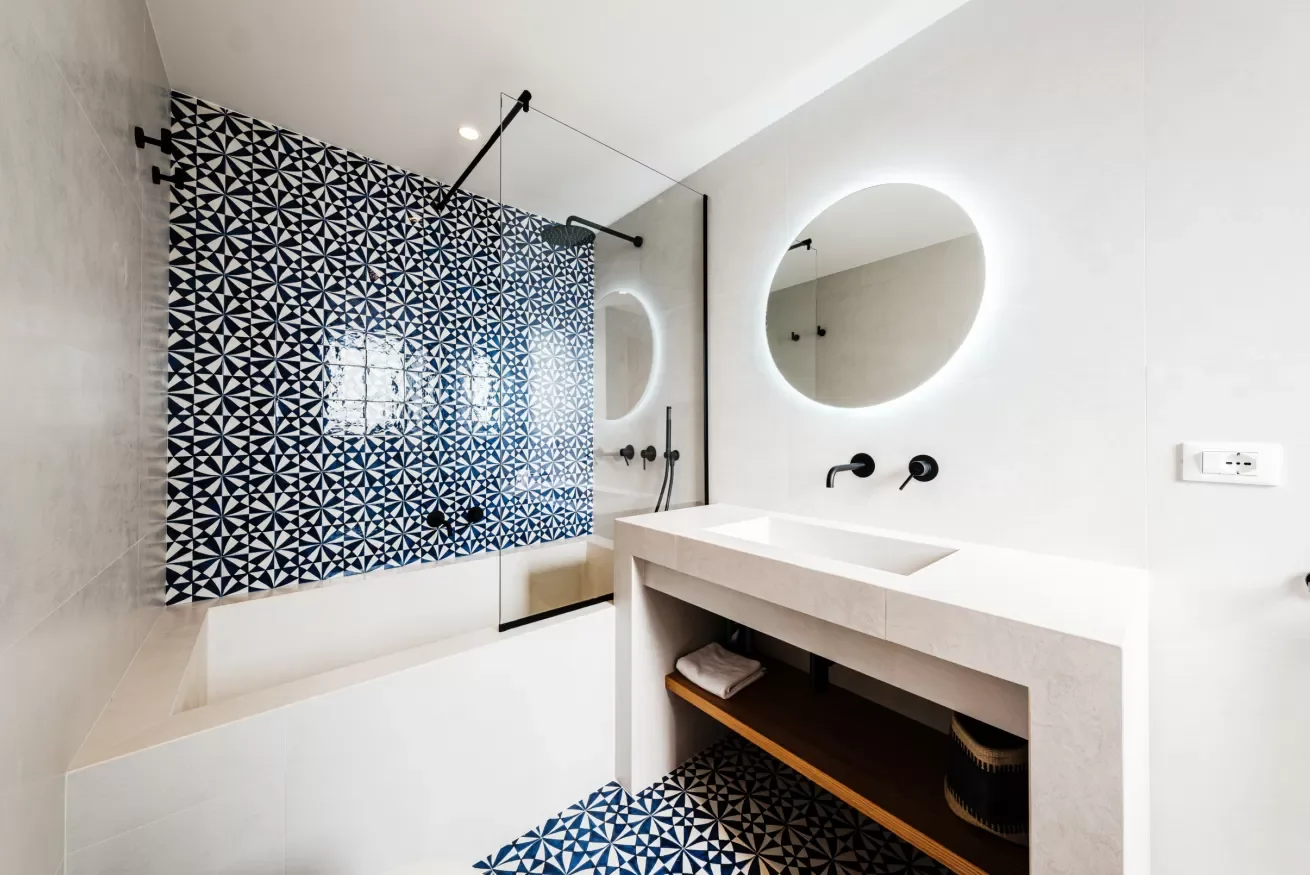 Modern bathroom with a bathtub, patterned shower curtain, round mirror with backlit LED, black fixtures, and a white vanity with a sink, towel, and storage underneath.