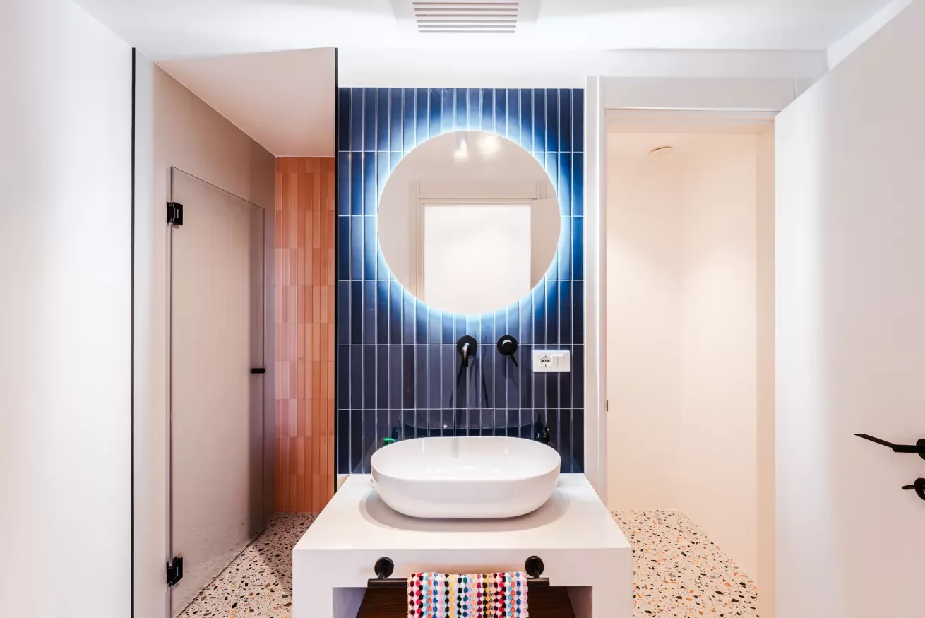 Modern bathroom with a round illuminated mirror, blue tiled accent wall, white vessel sink, terrazzo floor, and minimalist decor.