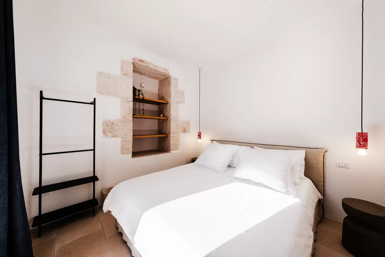 A minimalist bedroom with a white bed, beige headboard, black shelving unit, exposed stone wall niche with wooden shelves, and red pendant lights hanging on either side of the bed.
