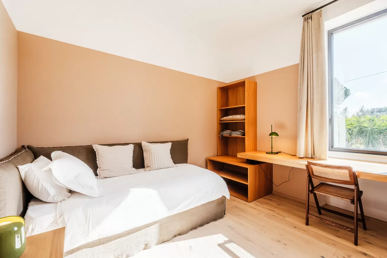 A cozy bedroom with a bed, pillows, a wooden desk, a chair, a tall window with cream curtains, and a shelving unit, illuminated by natural light