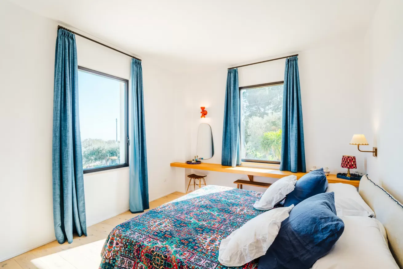 A bright bedroom with white walls, two windows with blue curtains, a bed with white and blue pillows, a colorful patterned bedspread, a wooden desk with a mirror and small stool, a wall-mounted lamp, and a small table with a red lamp.