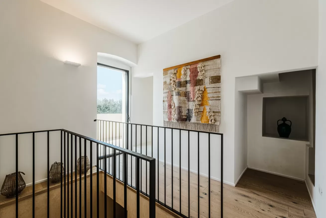 Interior view of a bright staircase landing with white walls, a window with a balcony door, a wall hanging, and a small niche with a decorative vase.