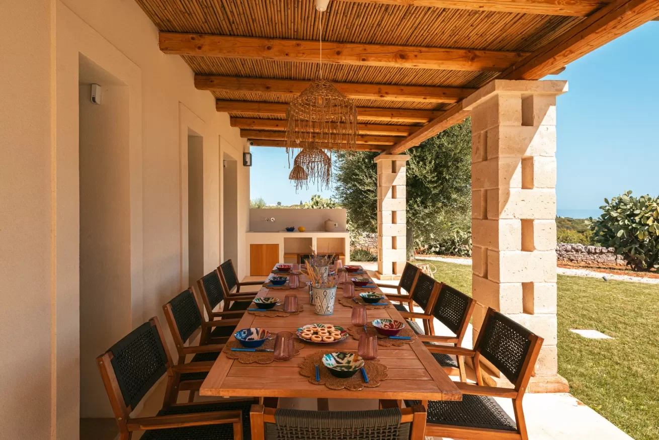 Outdoor dining table set under a wooden pergola in a garden with a grassy yard, large trees, and a stone wall in the background.