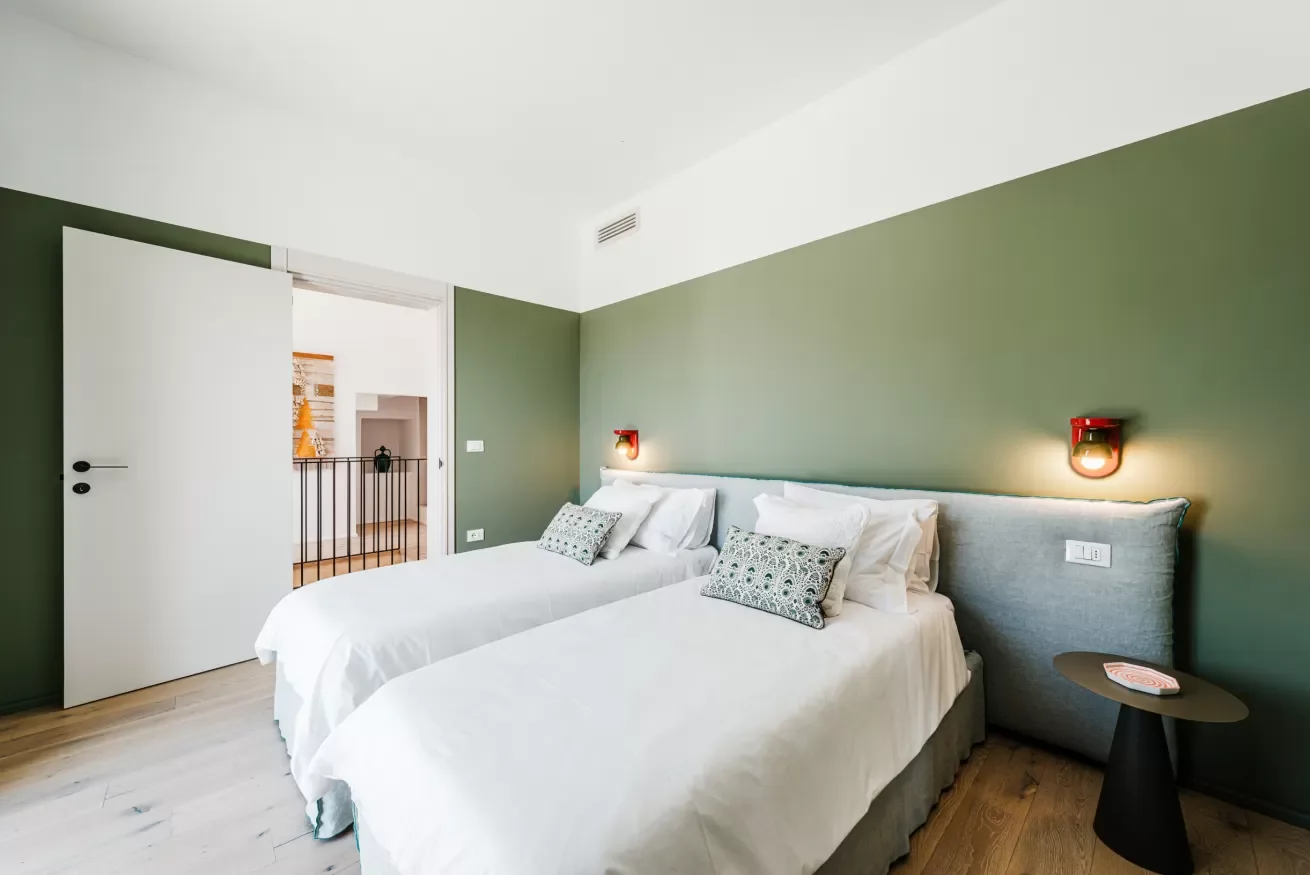 A bedroom with two twin beds, green accent wall, white bedding, patterned pillows, wall-mounted lamps, and a small black side table with a coaster.