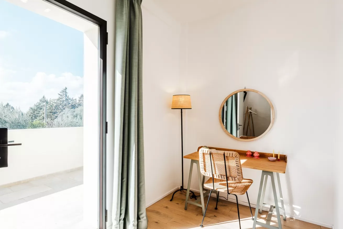 A minimalist room with a wooden desk, wicker chair, round mirror, standing lamp, and open door leading to a balcony with trees outside.