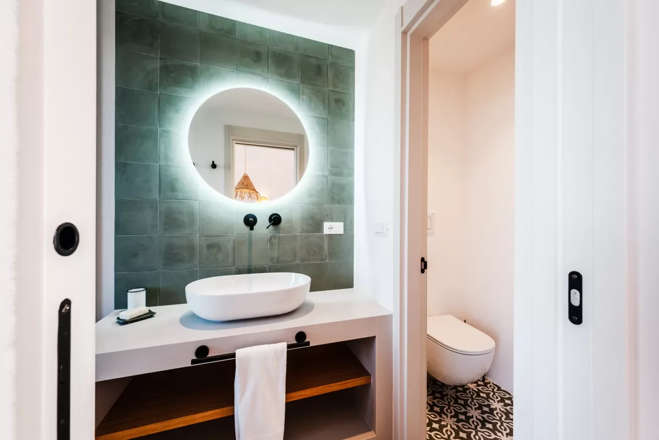 A modern bathroom with a circular backlit mirror, green tiled wall, white countertop, vessel sink, black fixtures, and a toilet in a separate enclosed space with patterned floor tiles.