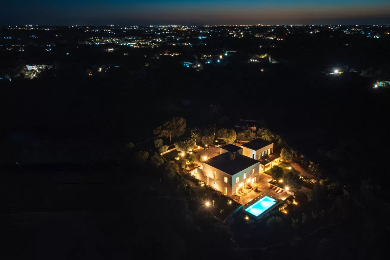 An aerial night view of a house with outdoor pool, lit by exterior lights, surrounded by trees, with a cityscape in the distance illuminated by lights.
