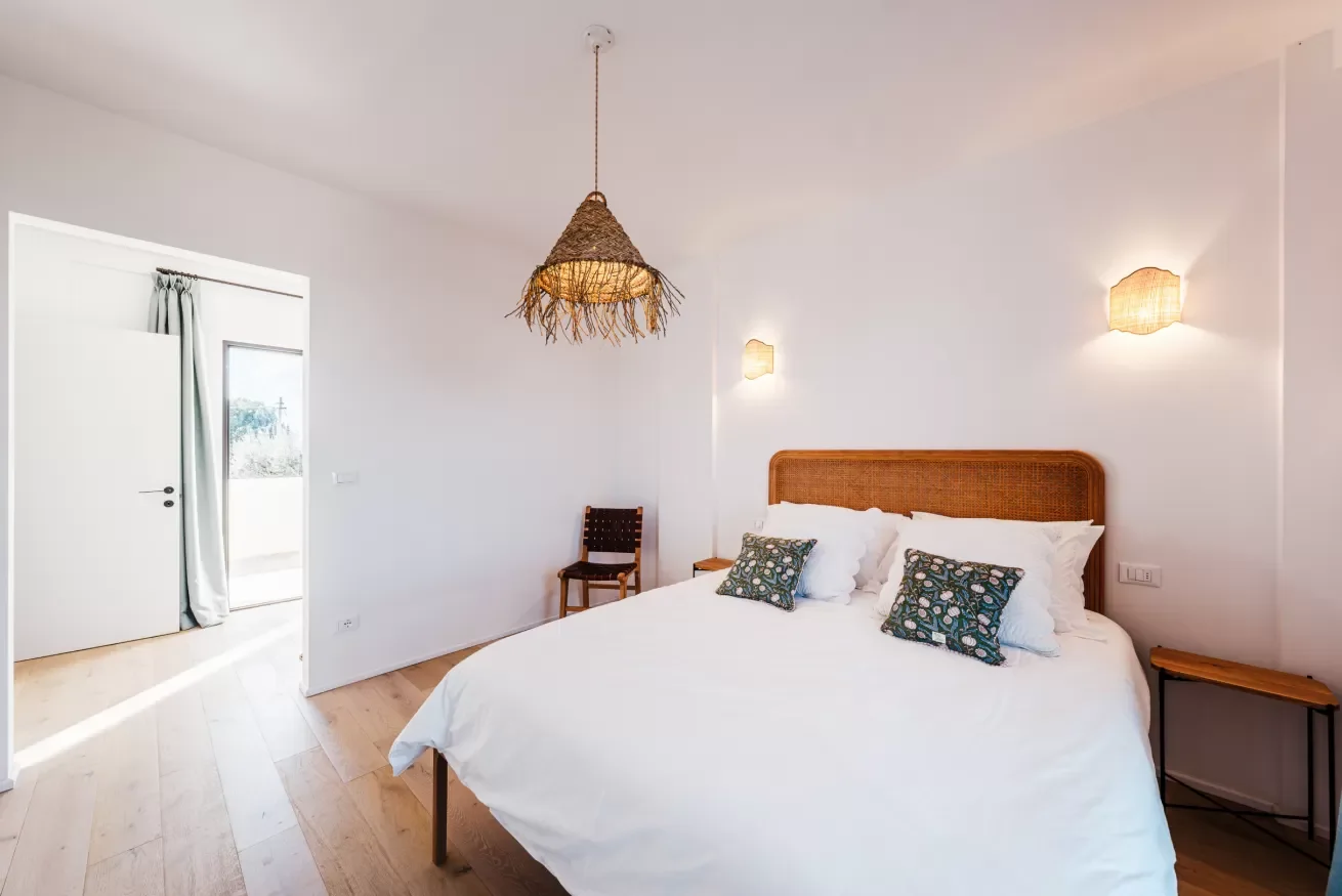 A bedroom with a large bed, wooden headboard, and decorative pillows, illuminated by wall sconces and a hanging wicker lamp, with a door leading outside.