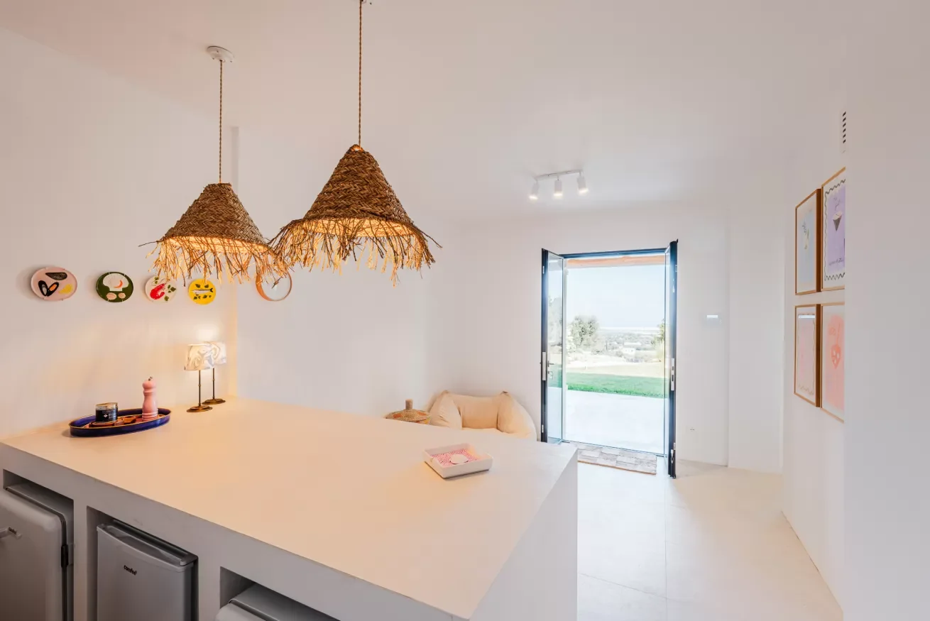 Bright room with a large white kitchen island, three hanging straw pendant lights, colorful wall art, a door opening to a green landscape, and a small sitting area with a beige cushion.