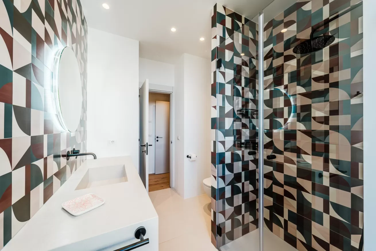 Modern bathroom with geometric patterned tiles on the walls in black, white, beige, and brown. Features a glass shower door, a white vanity with a black faucet, and a round mirror with LED lighting. White walls, ceiling with recessed lighting, and a 