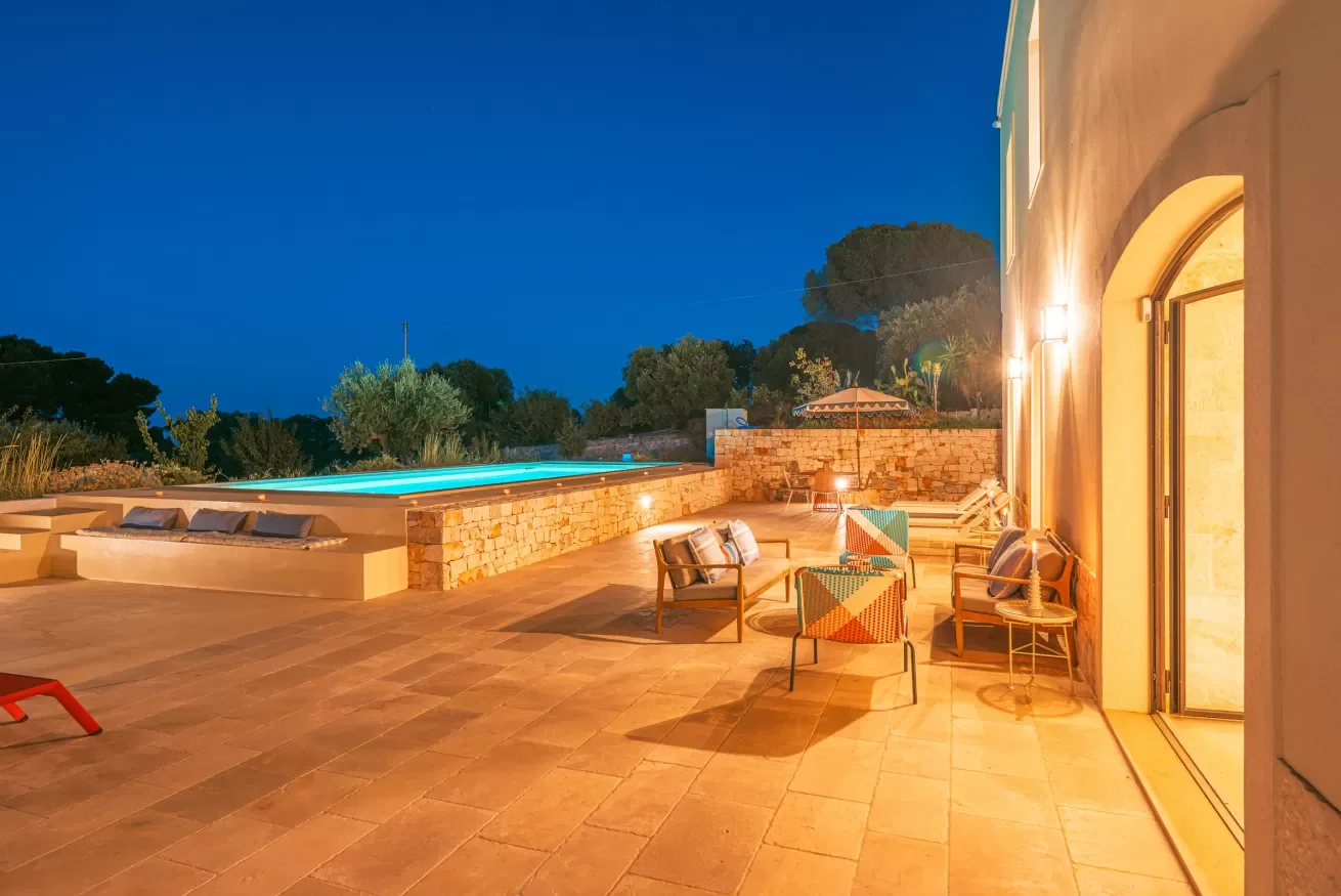 Outdoor patio at night with a lit swimming pool, seating area with chairs and cushions, and a stone wall, next to a house with illuminated walls.