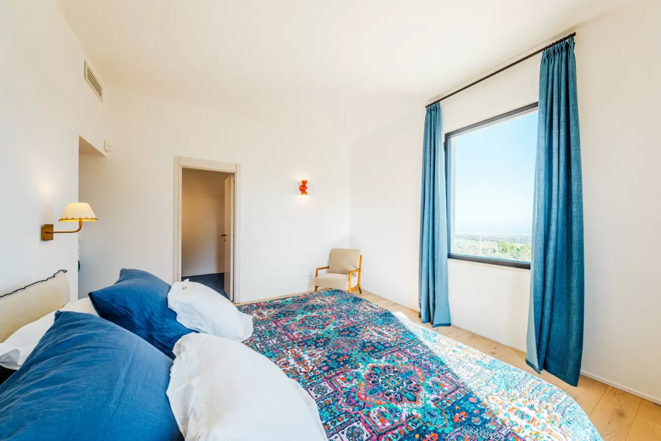 A bright hotel room with a large bed featuring blue pillows and bedding, a colorful patterned rug, a window with blue curtains showing a clear sky and distant landscape, a beige armchair by the window, and white walls with minimal decor.