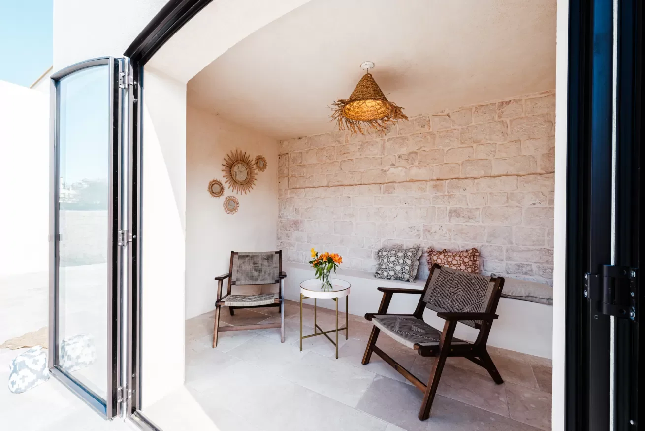 A cozy indoor seating area with two wooden chairs, a small round table with a vase of flowers, decorative pillows, a textured brick wall, and a woven pendant light.