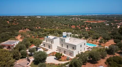Modern white house with multiple levels and flat roofs, surrounded by trees in a rural area with a distant ocean view