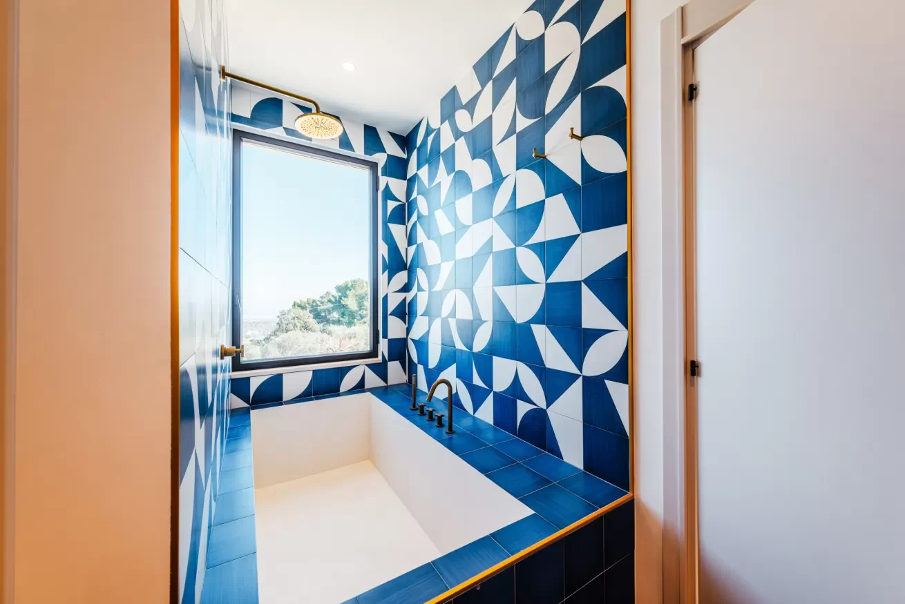 Bathroom with a built-in bathtub, blue geometric patterned tiles, a large window, and a gold showerhead.