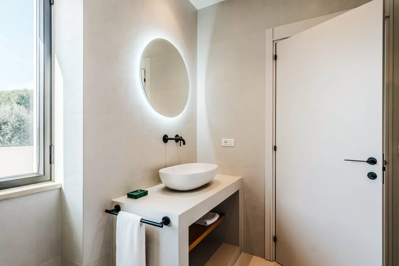 Modern bathroom with beige tiled walls, a white countertop with a vessel sink, a circular mirror with backlighting, a black wall-mounted faucet, a towel rack with a white towel, a small green tray on the counter, and a window showing trees outside.