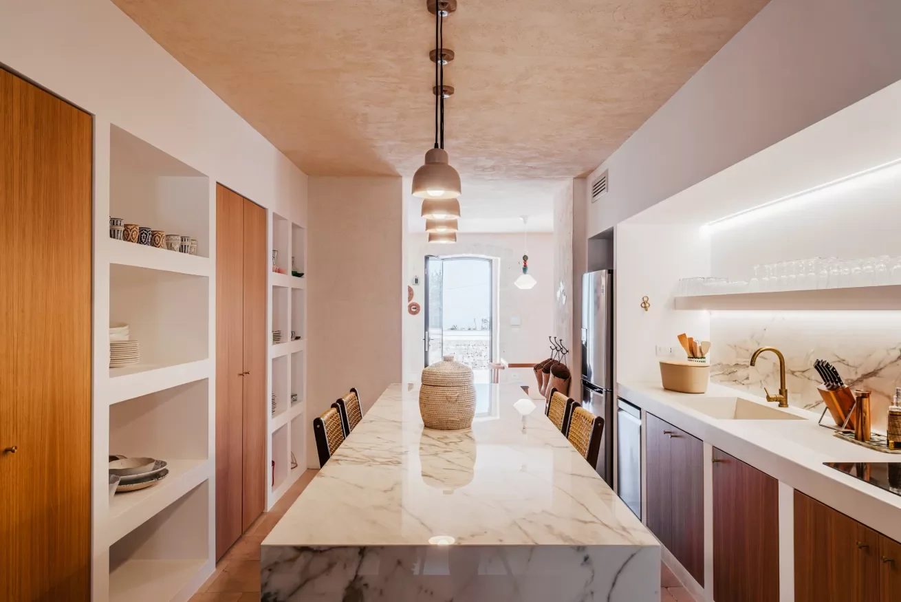 Modern kitchen with a marble island, wooden cabinets, open shelving, and a view to a bright outdoor space.