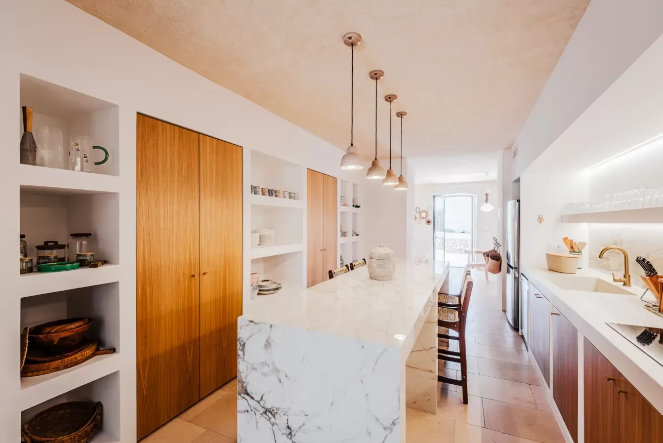 Bright modern kitchen with wooden cabinets, white shelves, and marble countertops, featuring pendant lighting and a view of a balcony through a glass door.