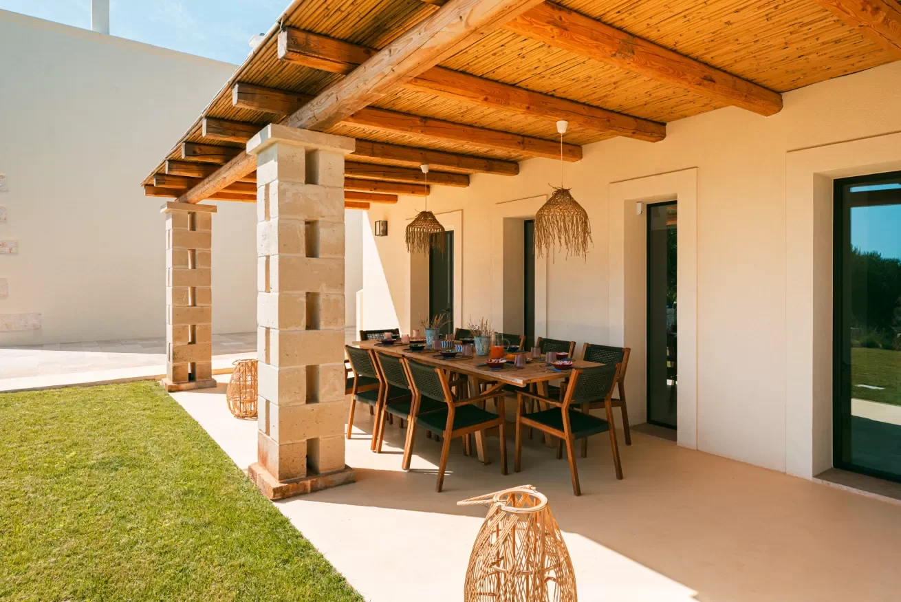 Outdoor covered patio with wooden dining table and chairs, hanging wicker light fixtures, stone columns, and a grassy yard area.