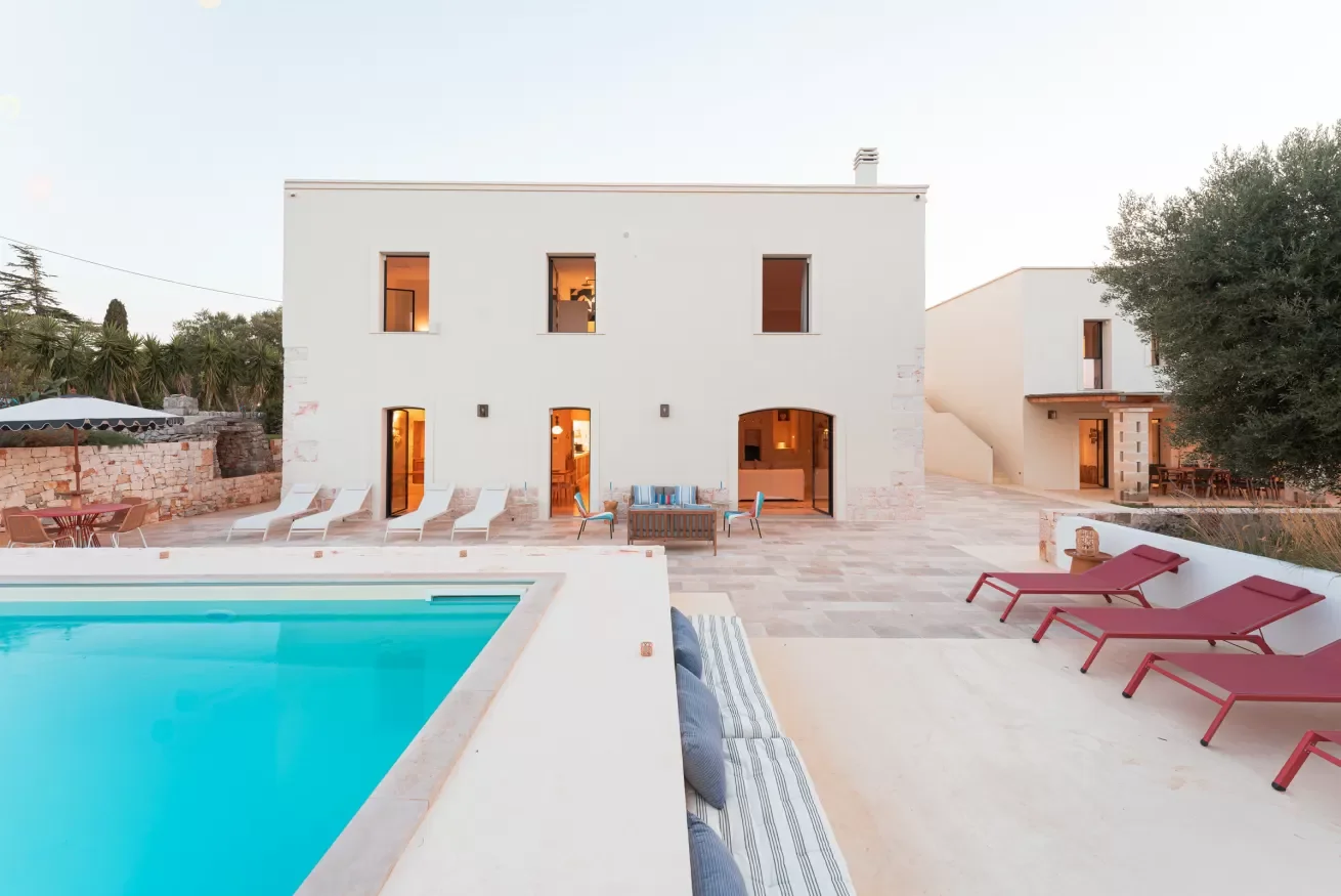 Modern white house with multiple windows and sliding doors, pool in foreground, and outdoor seating and lounge chairs on patio.