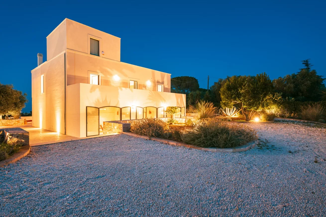 A modern, multi-story house illuminated at night with exterior lighting, surrounded by desert landscaping and sparse trees.