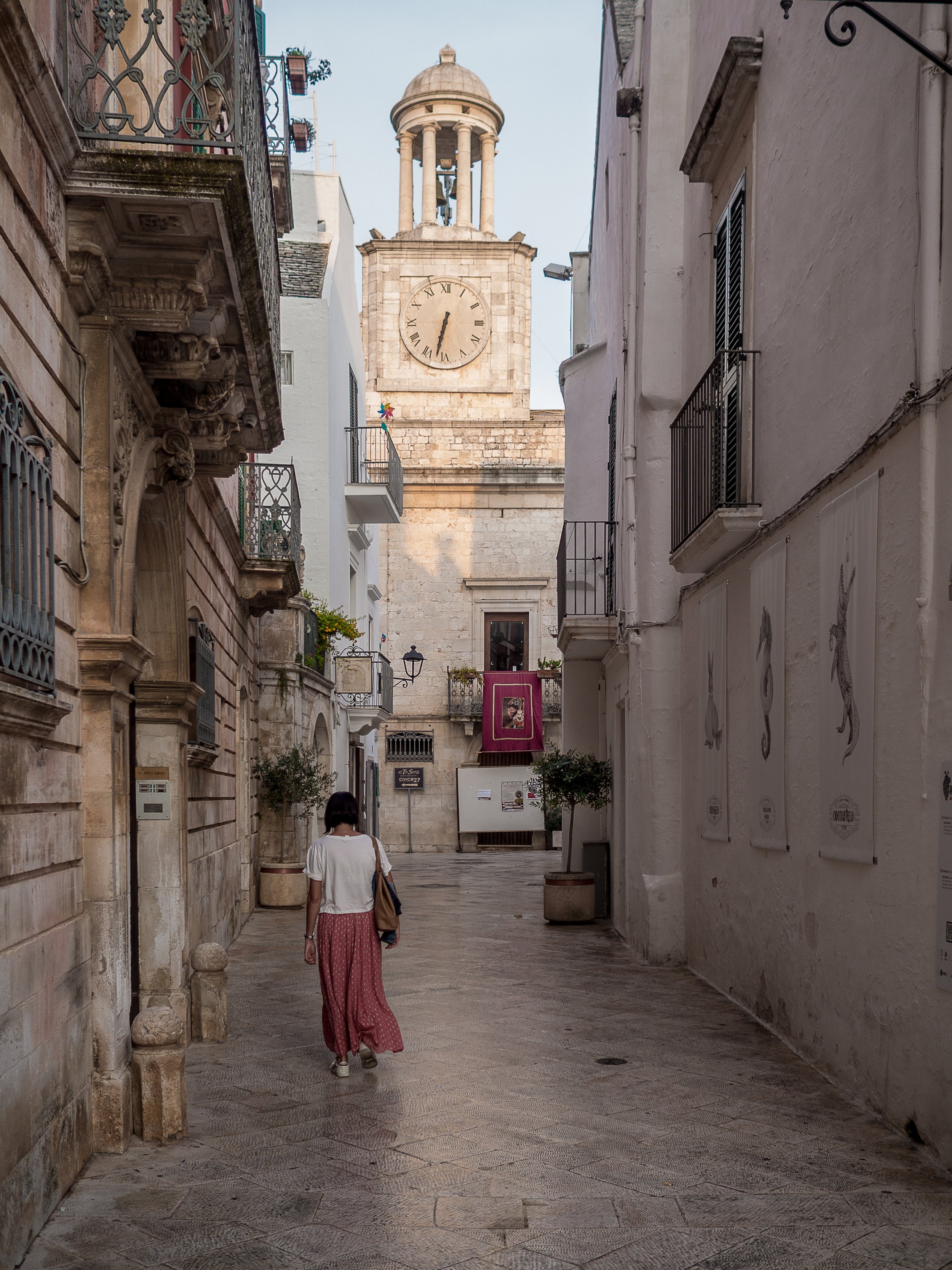 A woman walking down a narrow European street with old buildings and balconies, with a clock tower in the background showing 4:20.