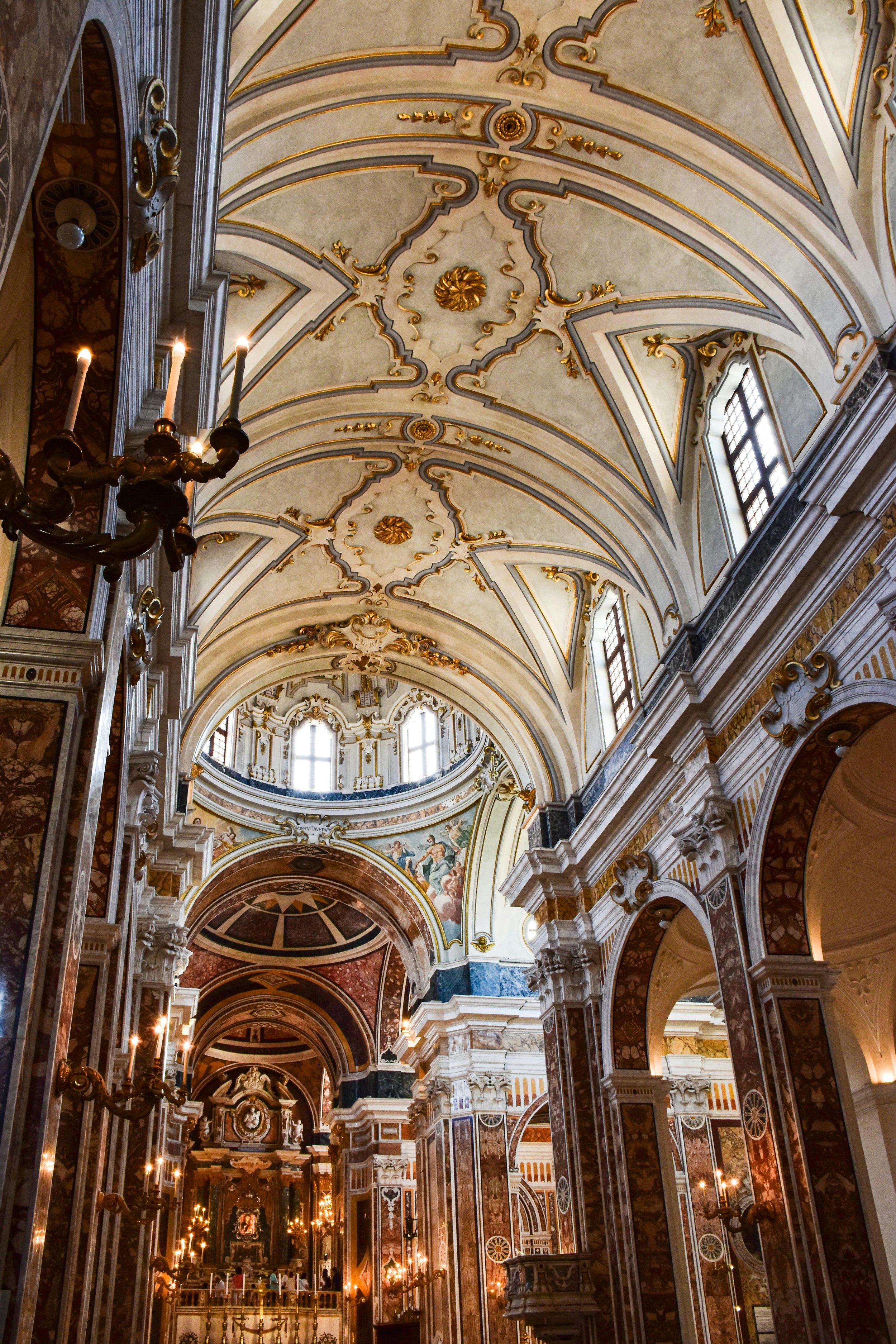 Interior view of a Baroque church or cathedral with ornate gold and marble detailing, arched ceilings, stained glass windows, and vintage wall sconces.