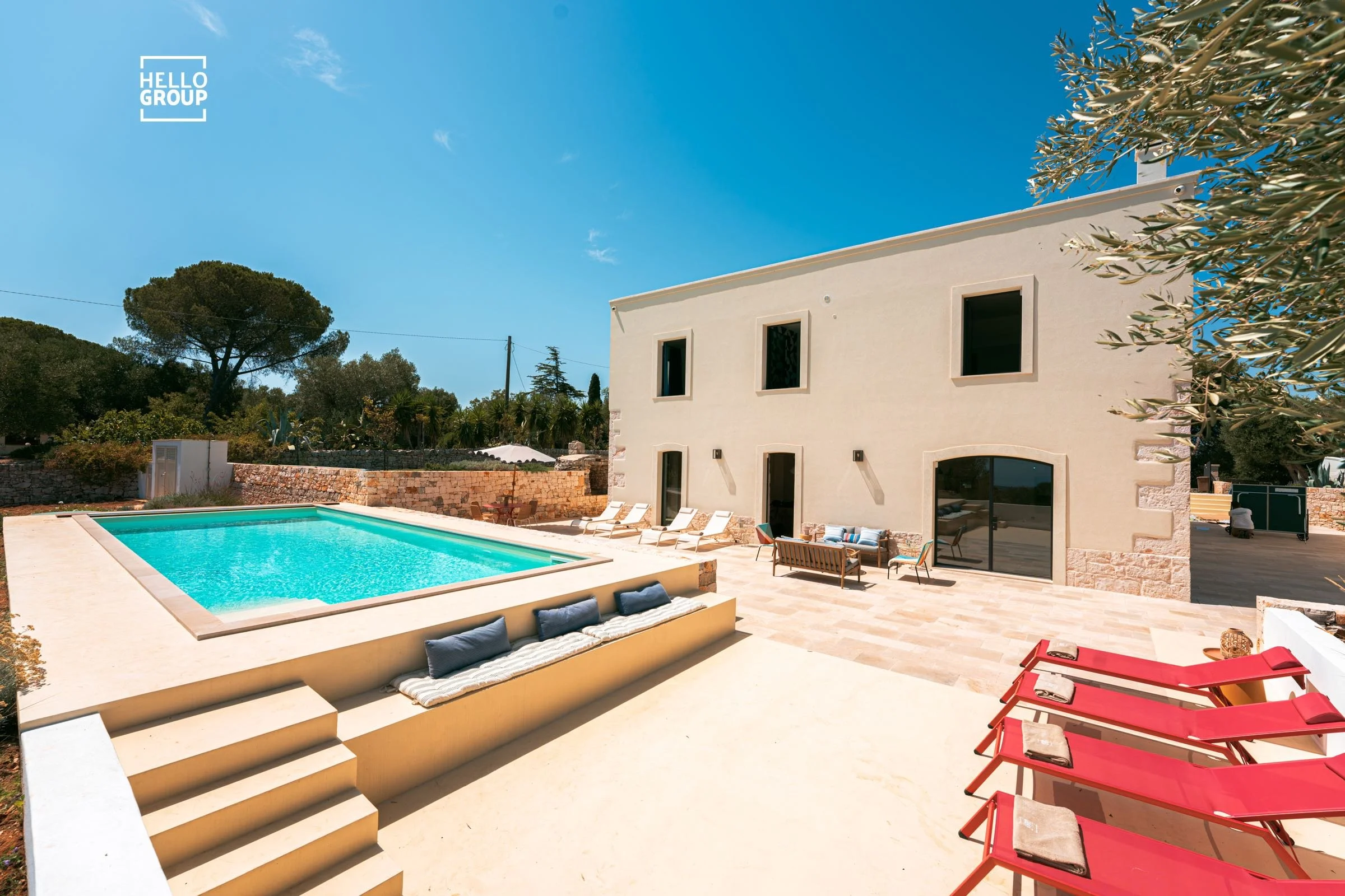 A modern house with a swimming pool surrounded by a patio with lounge chairs and outdoor furniture, set against a clear blue sky and trees.