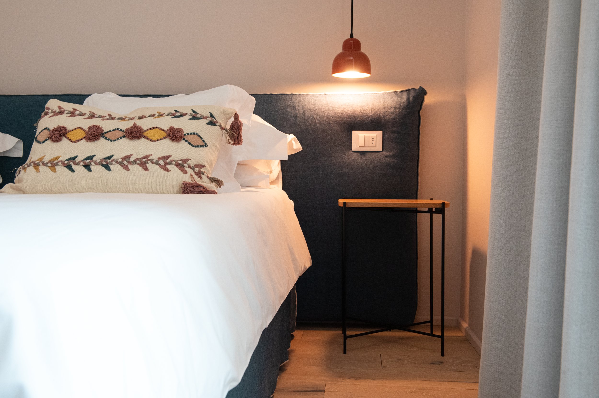 A modern bedroom corner with a bed featuring white linens and a decorative embroidered pillow. There is a wall-mounted lamp emitting warm light, a power outlet, and a minimalist bedside table. A curtain is partially visible on the right side.