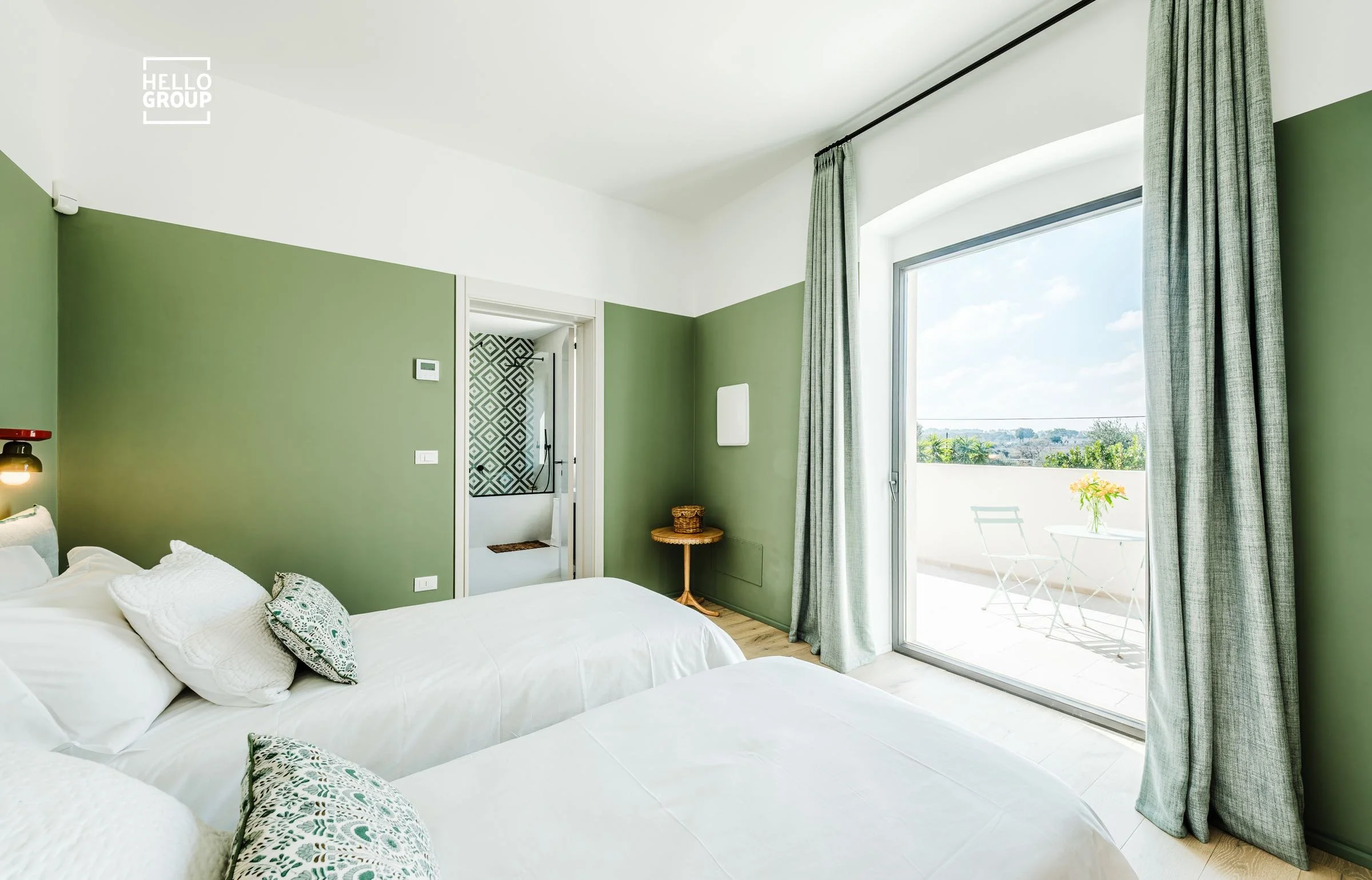 Bright hotel room with two beds, green accent walls, and a sliding glass door leading to a balcony with outdoor seating and a potted plant.