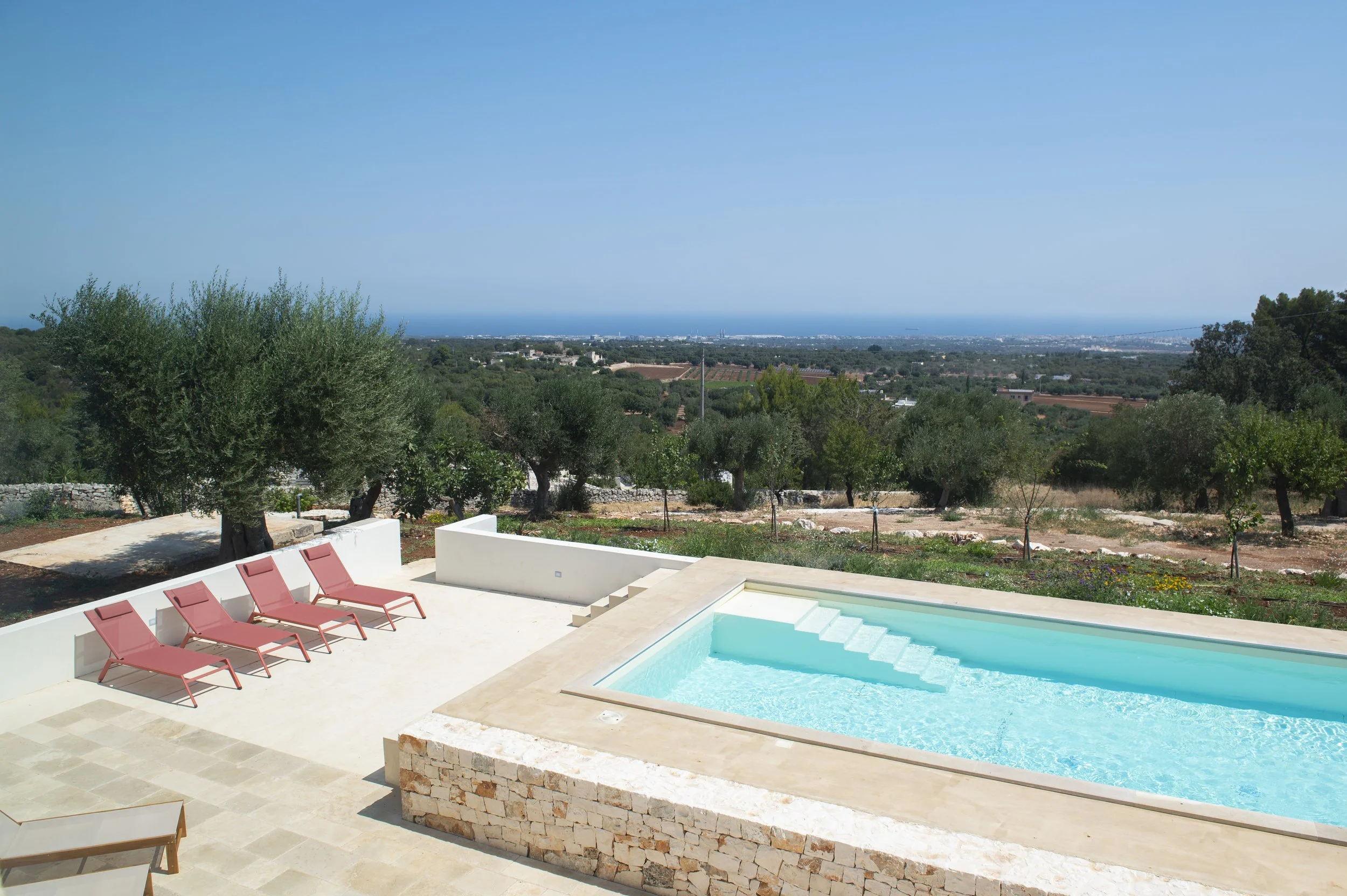 A swimming pool with clear blue water, surrounded by pink lounge chairs on a white patio, overlooking an expansive landscape of trees, fields, and a distant view of the ocean under a clear blue sky.