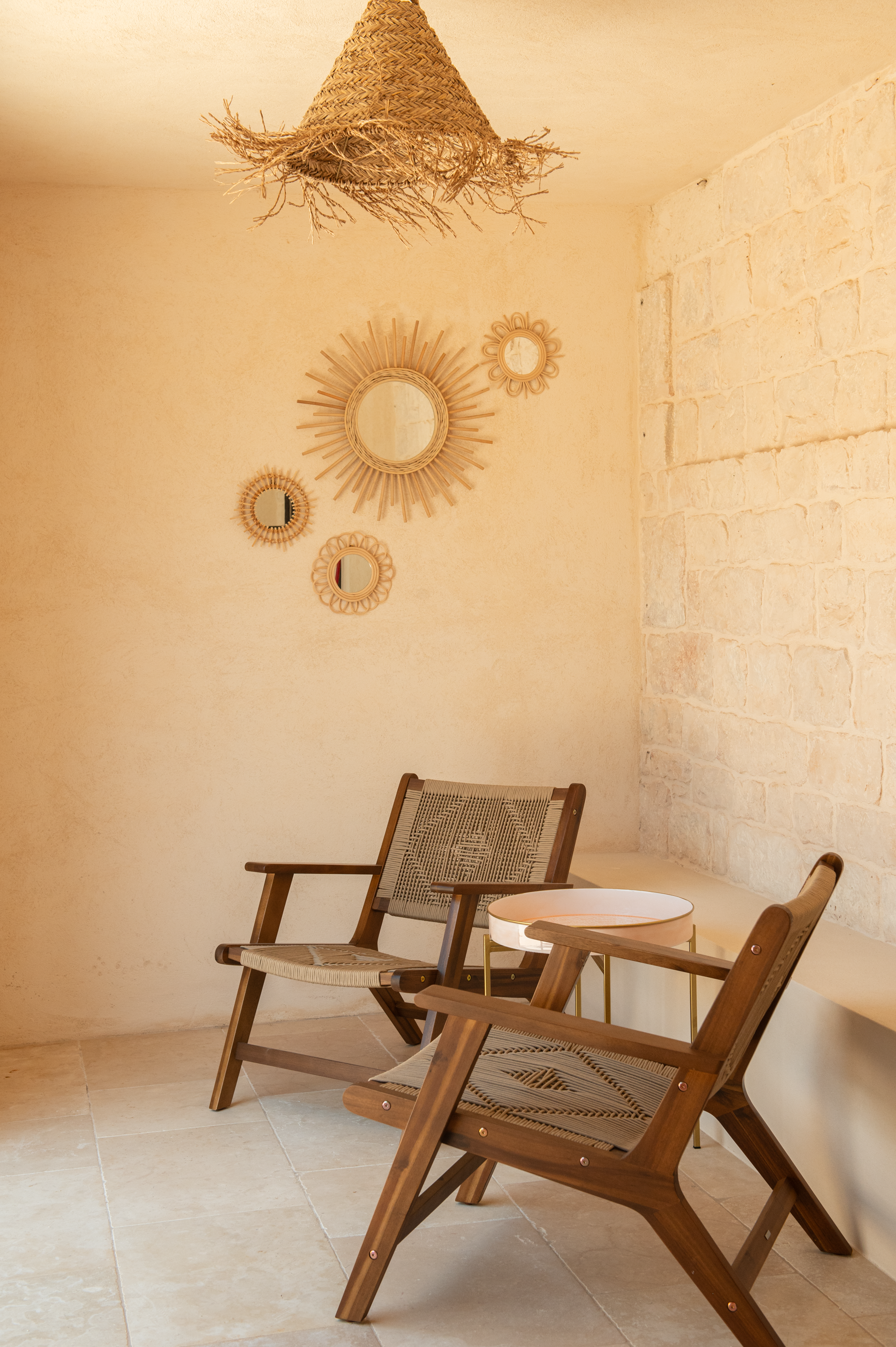 Cozy corner with two wooden chairs, a small side table, a textured white brick wall, and decorative round mirrors on the wall. A woven lampshade hangs from the ceiling.