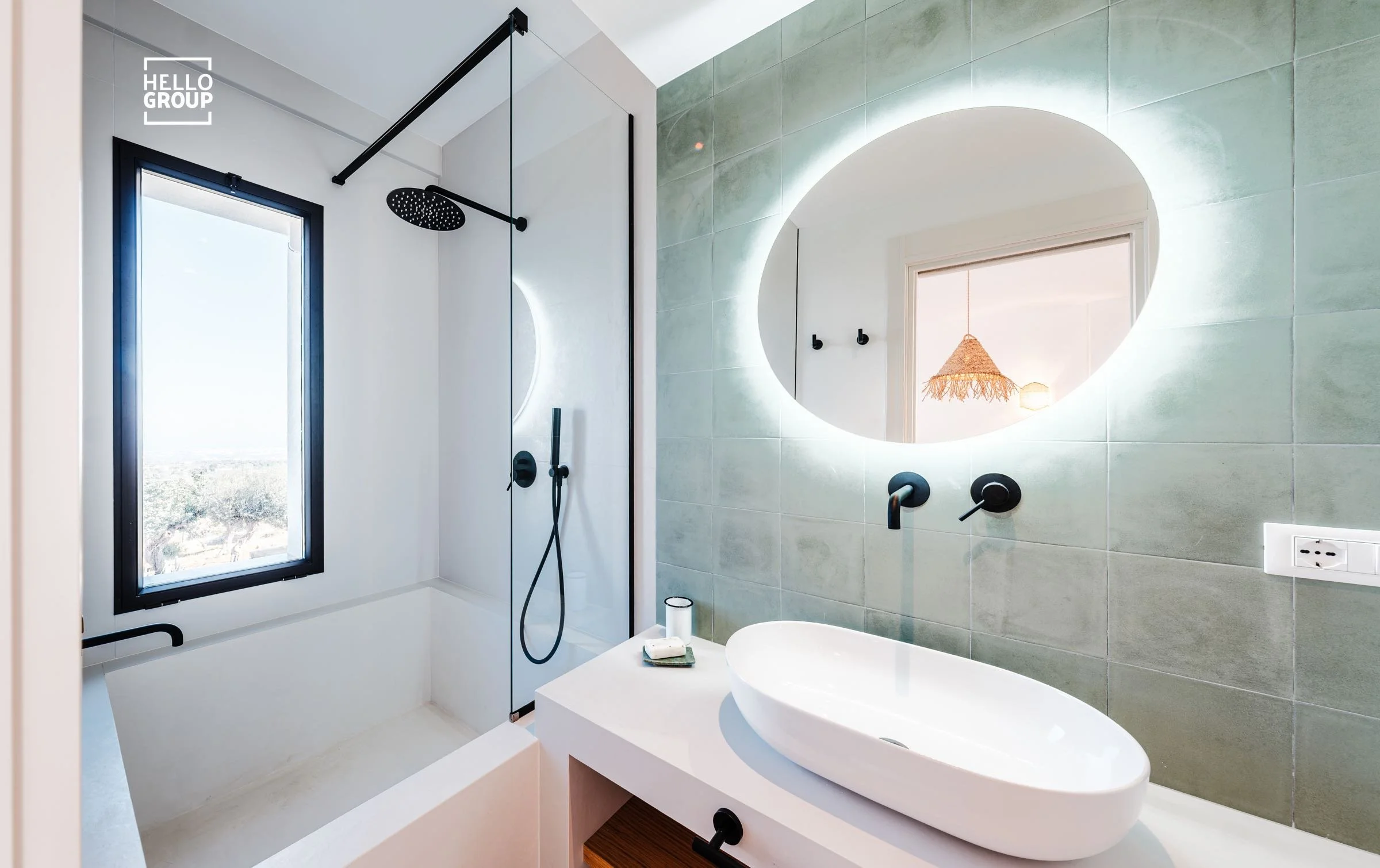 Modern bathroom with a walk-in shower, large round illuminated mirror, oval white vessel sink, black fixtures, and a window with a mountain view.