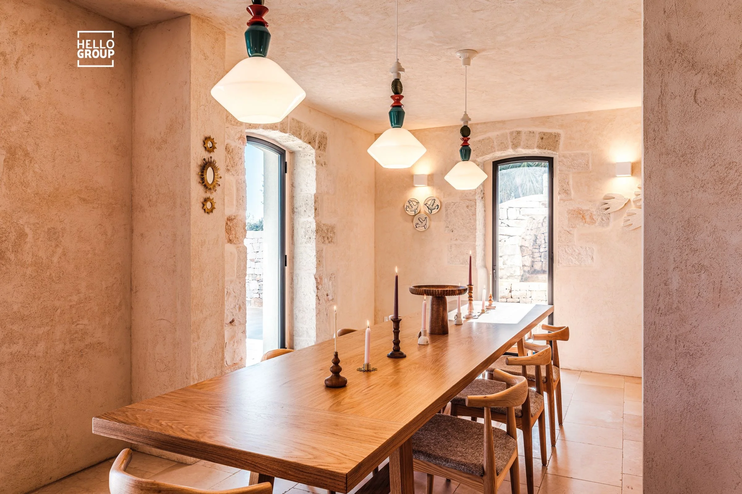 A cozy dining room with a long wooden table and six chairs. The room has stone walls, two tall windows, and hanging light fixtures. The table has candles and a decorative centerpiece.