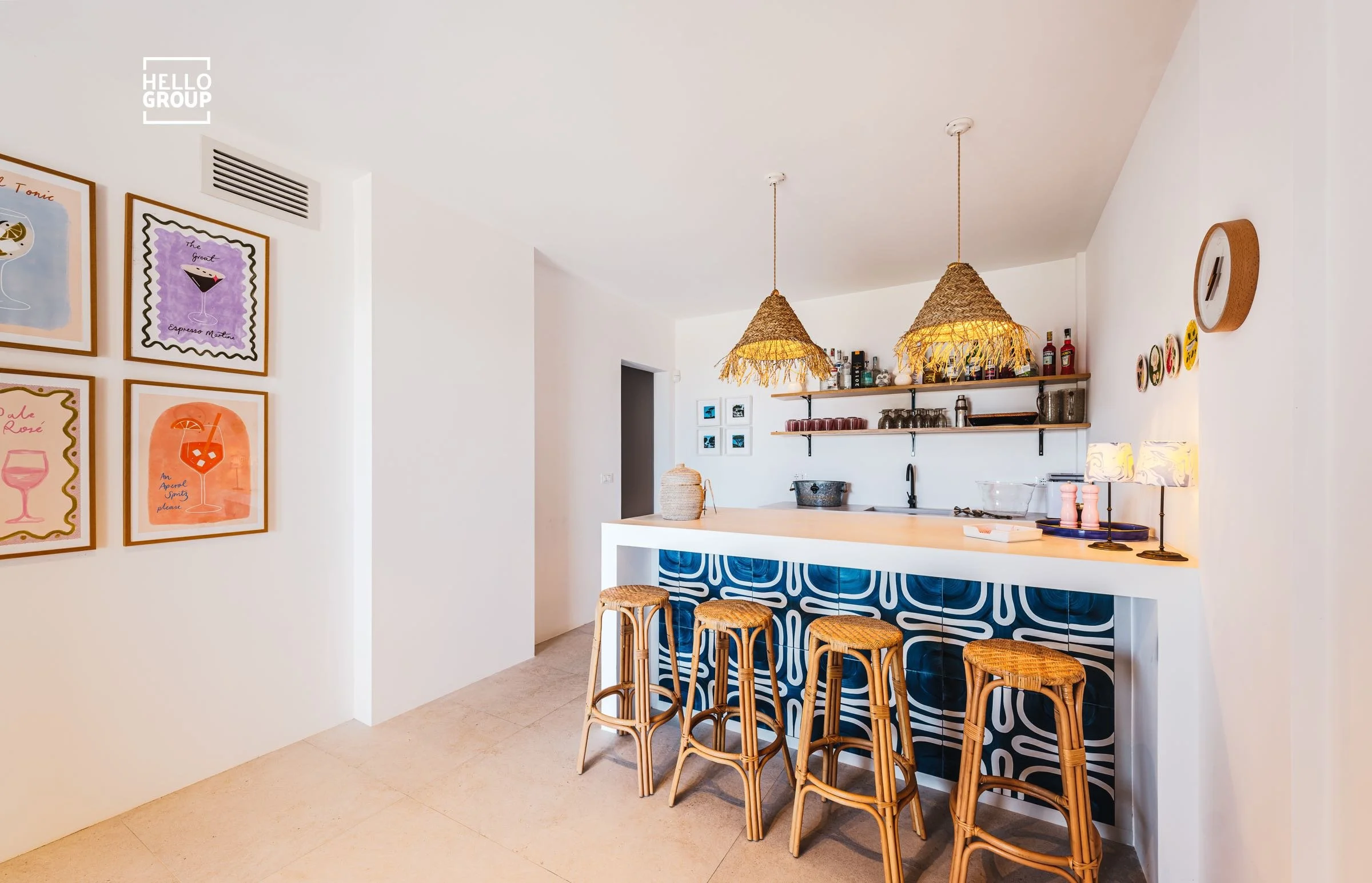 Bright kitchen area with white walls, a small bar with four rattan stools, and art frames on the left wall featuring colorful illustrations of drinks.