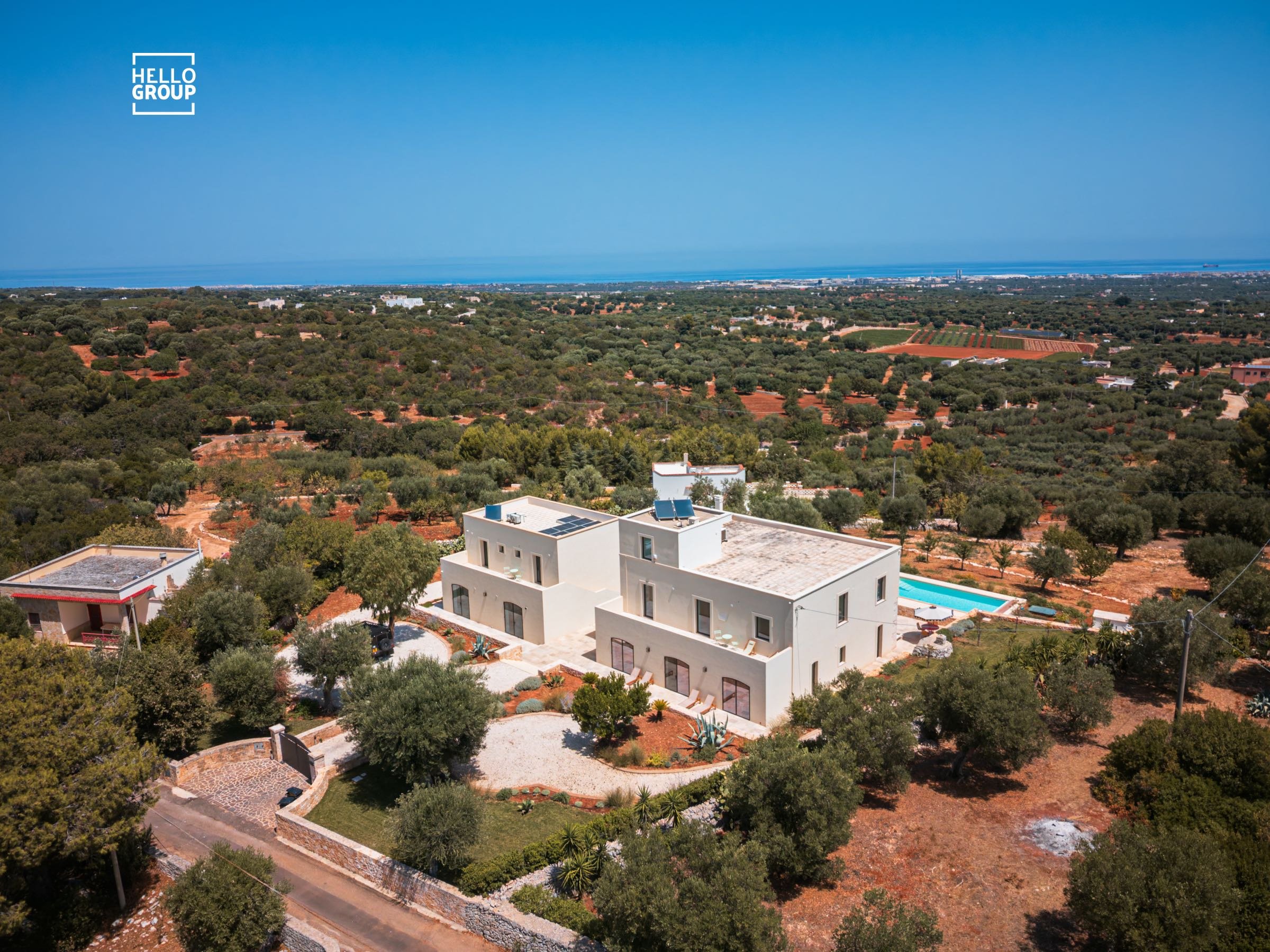 Aerial view of a large well designed and proportioned white house with a swimming pool, surrounded by trees and a rural landscape with fields and distant coastline under a clear blue sky.