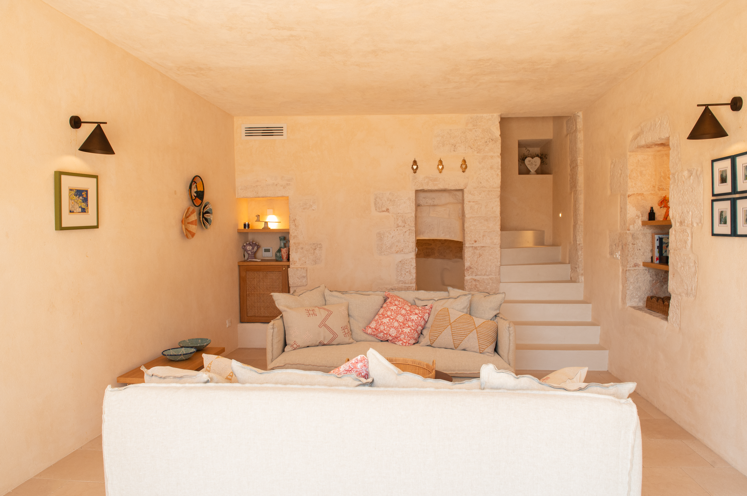 Living room with beige walls, a white sofa with decorative cushions, a small side table, wall-mounted lights, and built-in shelves with decorative items.
