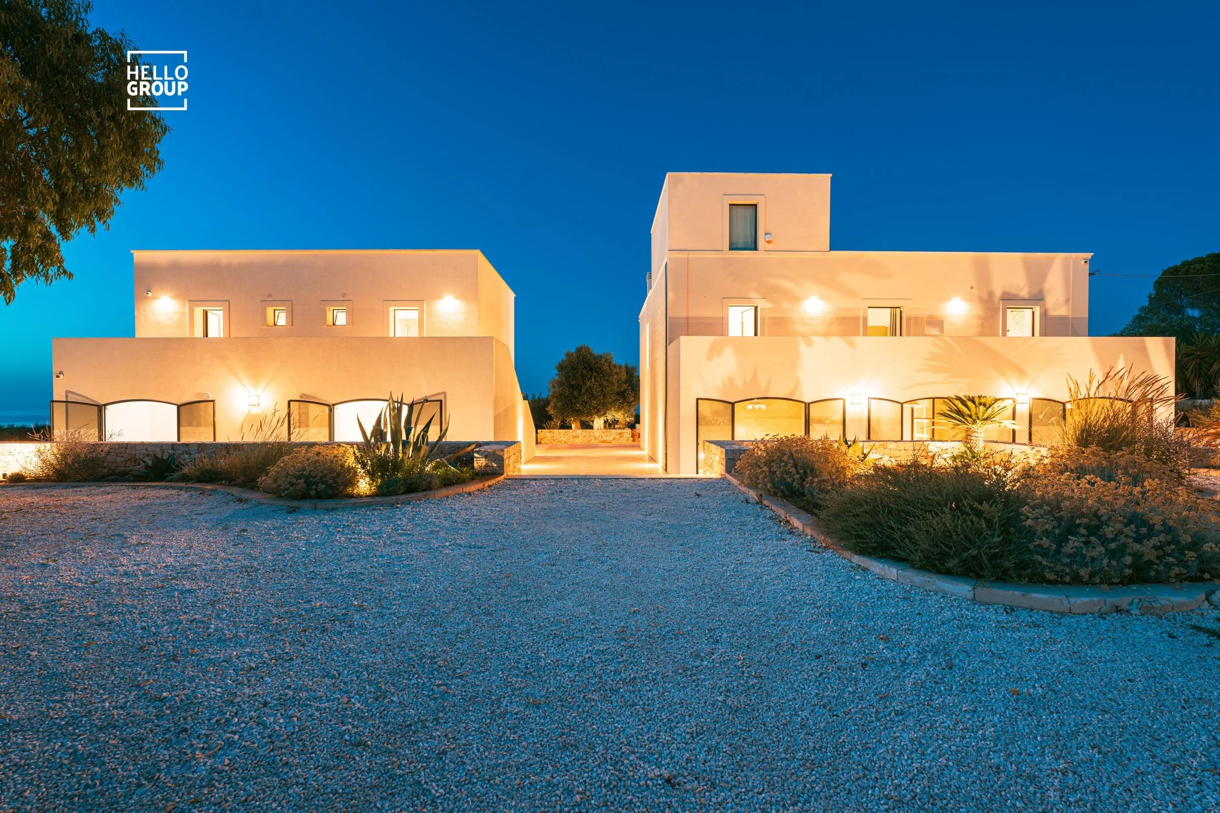 A classical pair of villas  illuminated at dusk, with plants and a gravel driveway in the foreground and an deep sky in the background.
