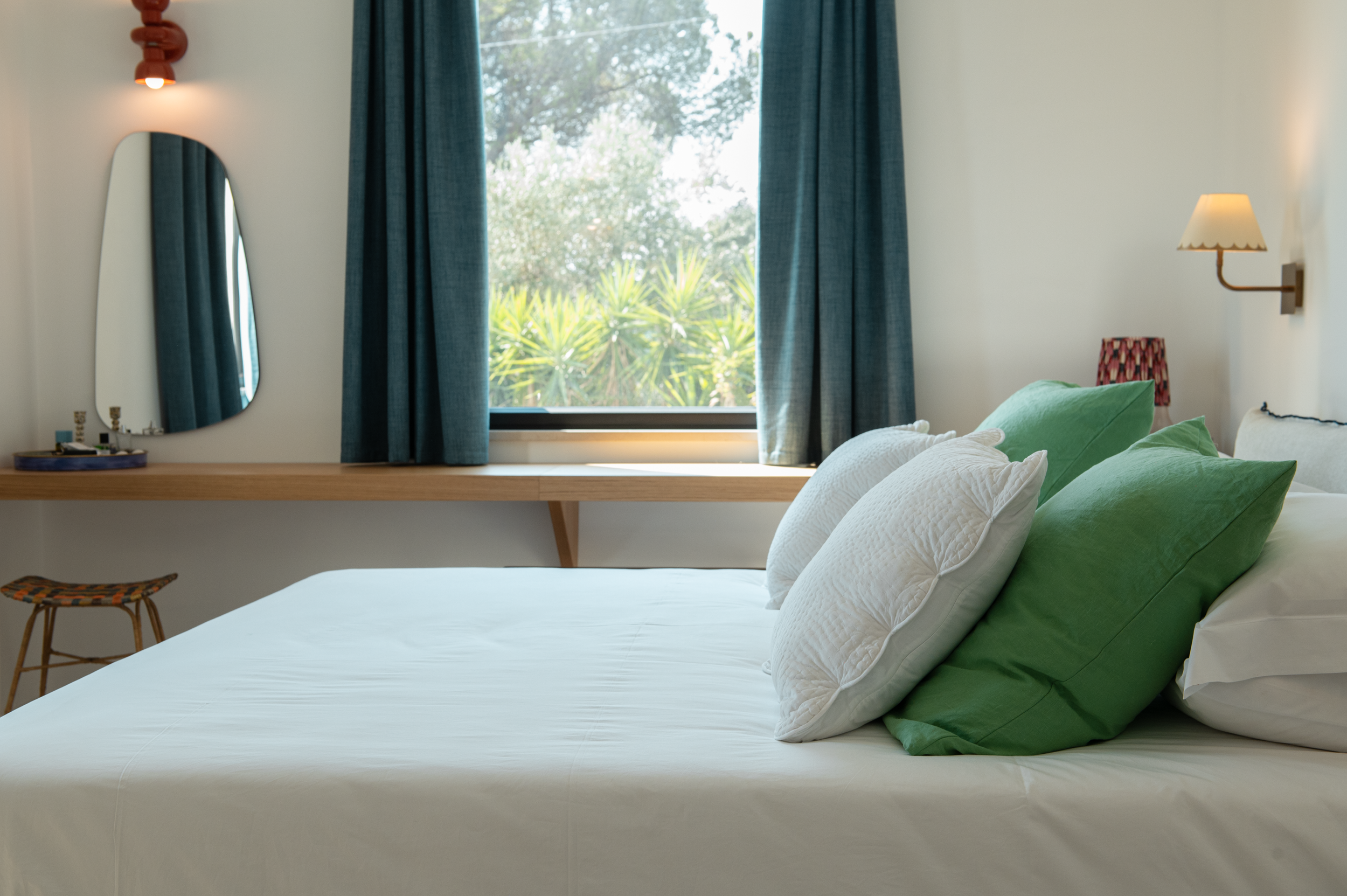 A bedroom with a bed decorated with green and white pillows, a window with blue curtains, a wooden desk with a mirror, and a small table with a patterned lampshade.