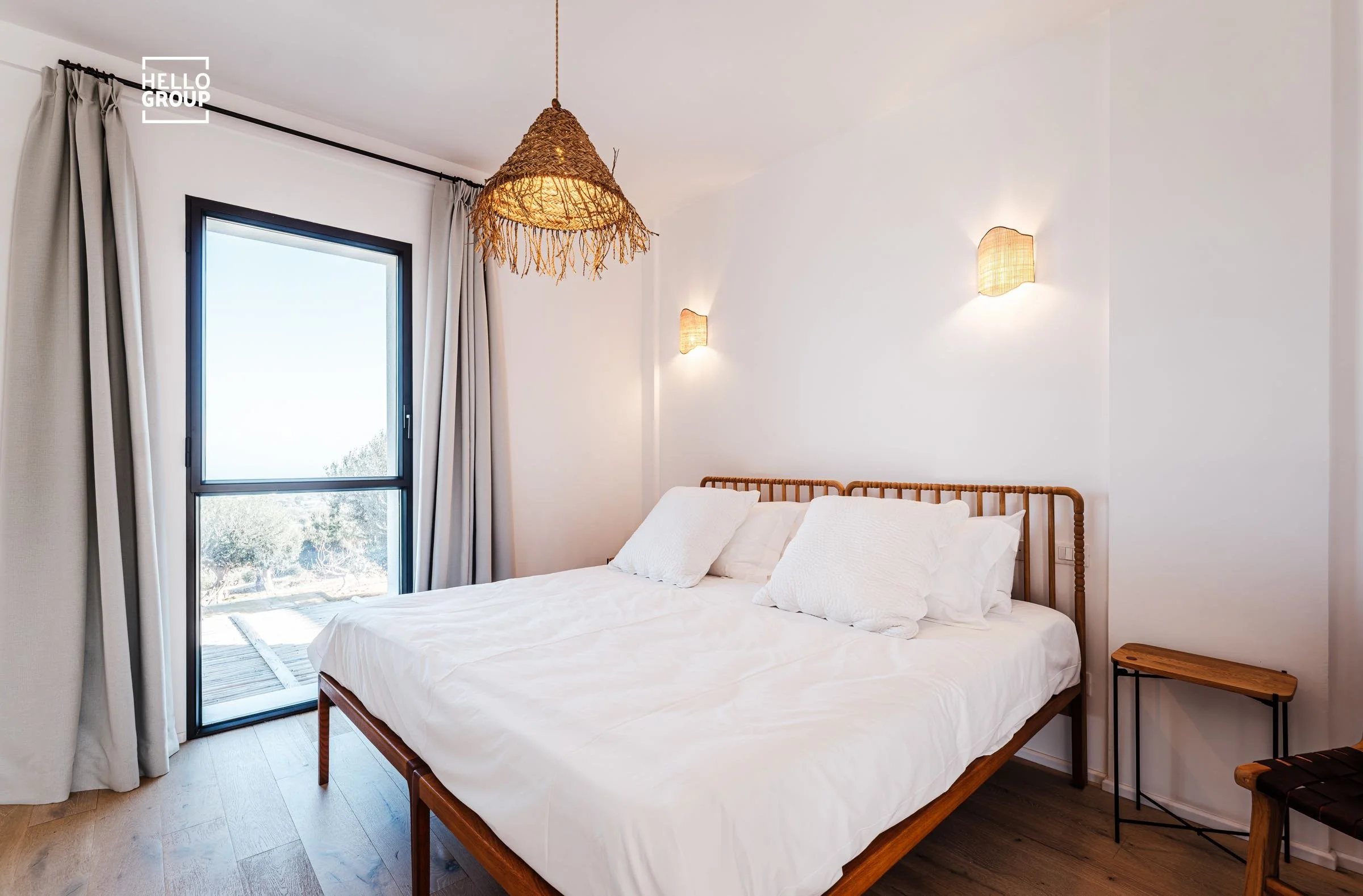 Bright bedroom with white walls, a large bed with white bedding and pillows, a bamboo headboard, a woven pendant light hanging from the ceiling, two wall-mounted lamps, a sliding glass door with beige curtains, a small wooden side table, and wooden f