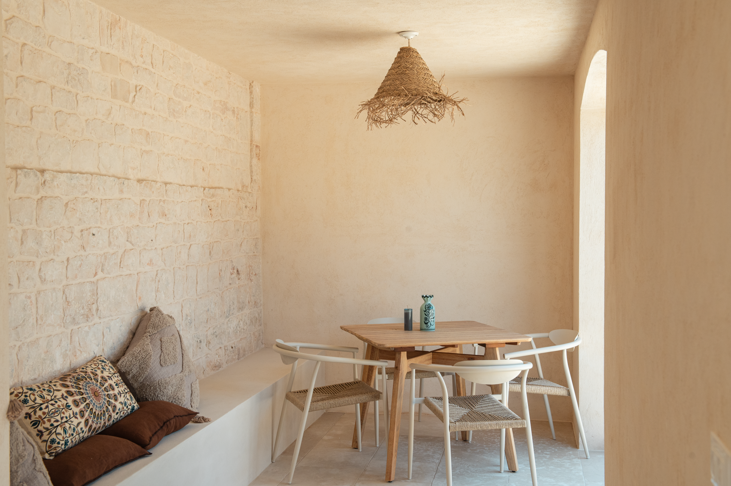 Dining area with beige walls, a wooden table with five chairs, decorative pillows on a built-in bench, and a woven cone-shaped light fixture hanging from the ceiling.