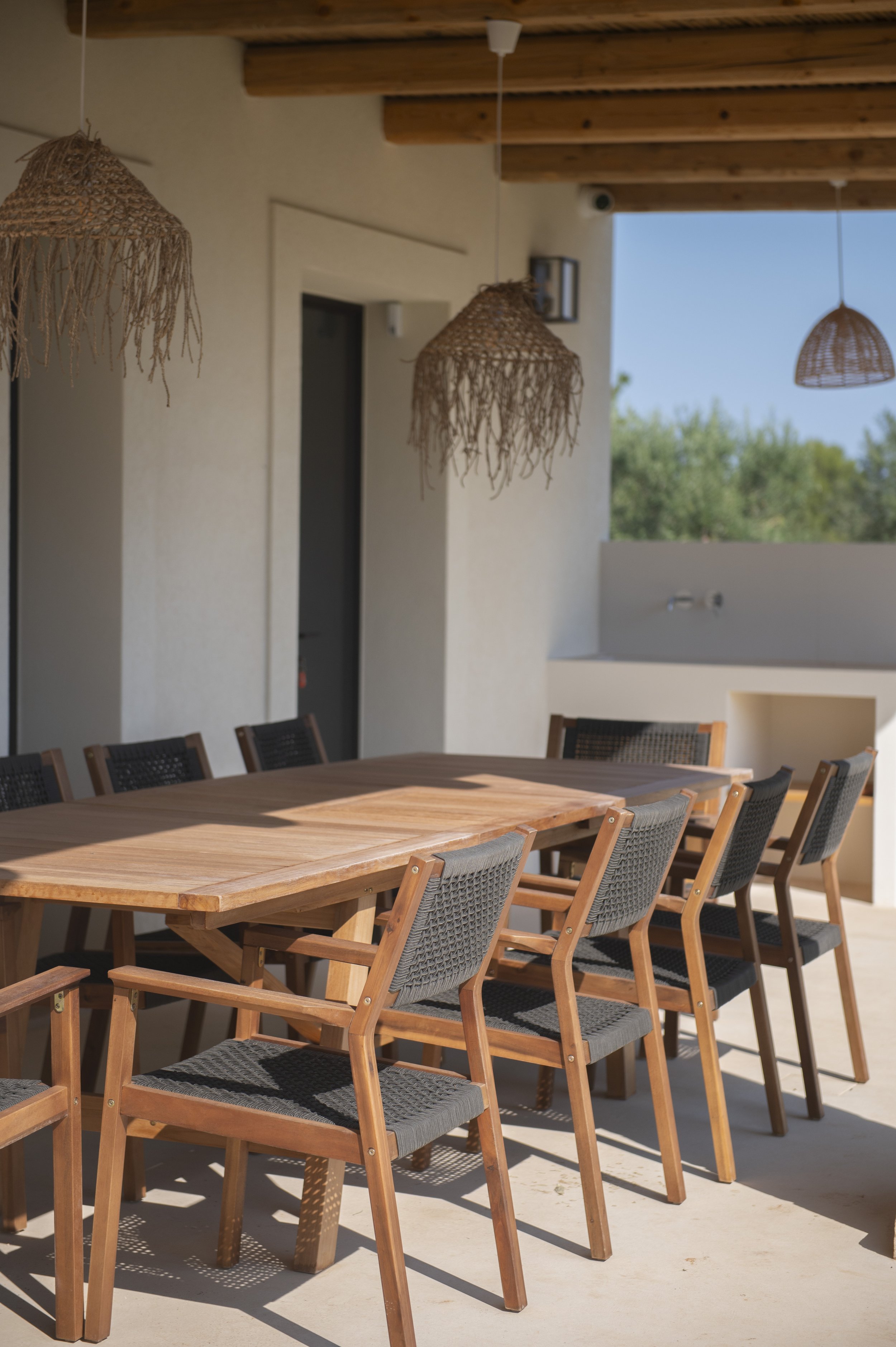 Outdoor dining area with a wooden table and matching chairs under wicker pendant lights, adjacent to a beige wall and outdoors with greenery visible in the background.