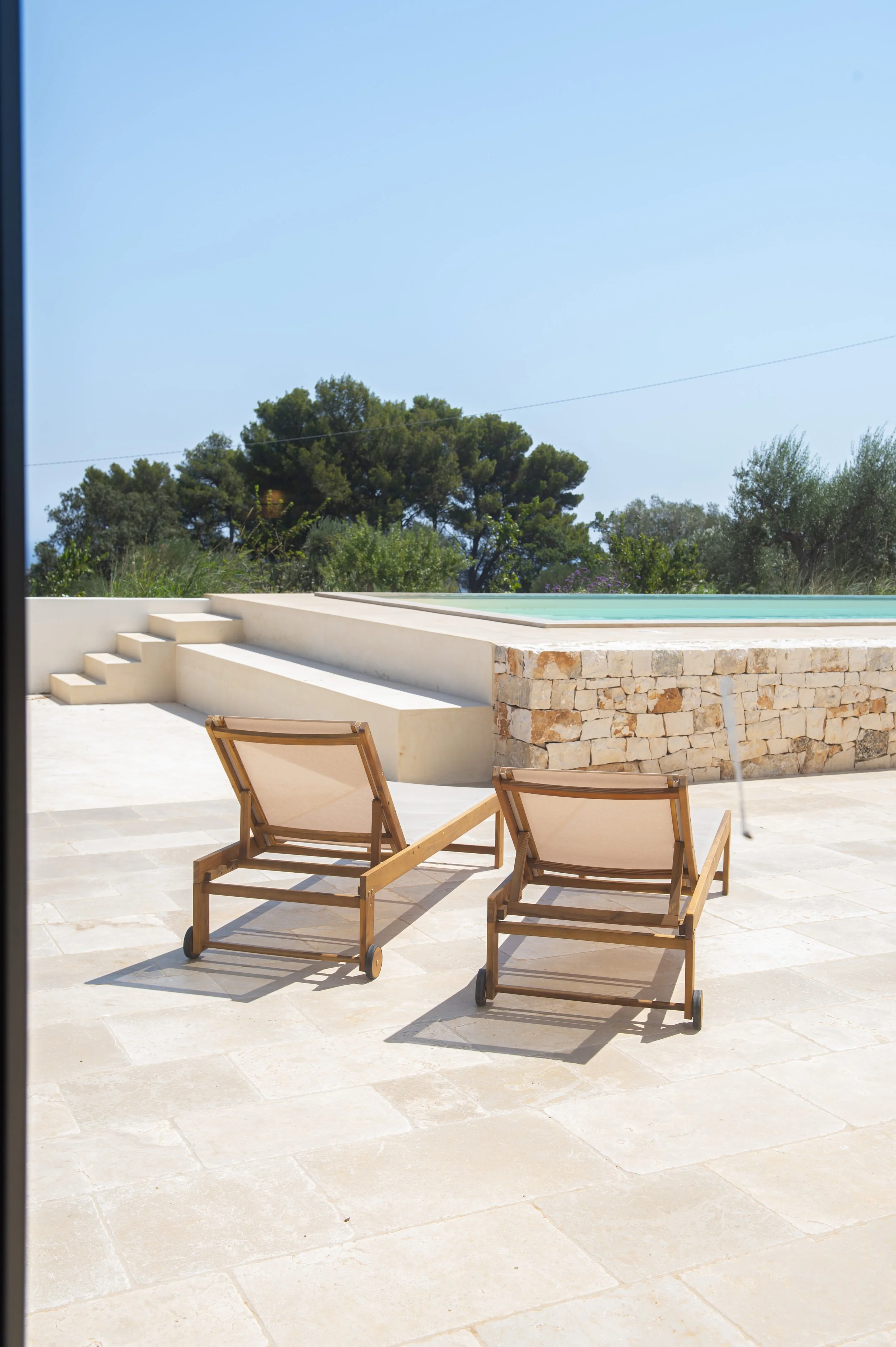 Two wooden lounge chairs with beige fabric seats on a light stone patio near a pool, with trees and a clear blue sky in the background.
