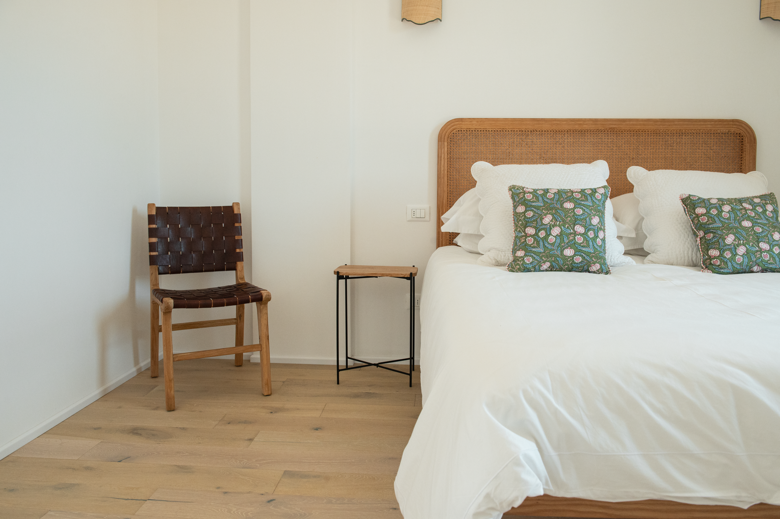 A bedroom with a wooden bed, white bedding, and colorful patterned pillows. There is a brown chair, a small side table, and a wall with a light switch.