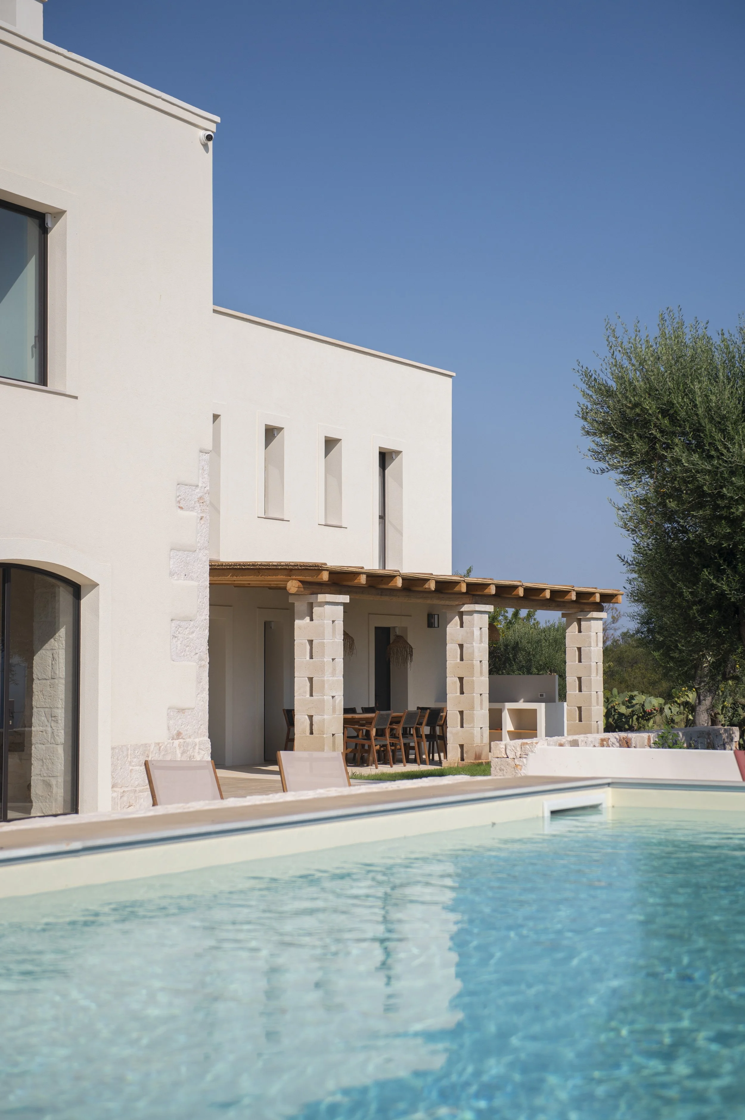 A modern house with a white exterior, a covered patio with wooden beams, outdoor dining furniture, and a swimming pool in the foreground, under a clear blue sky.