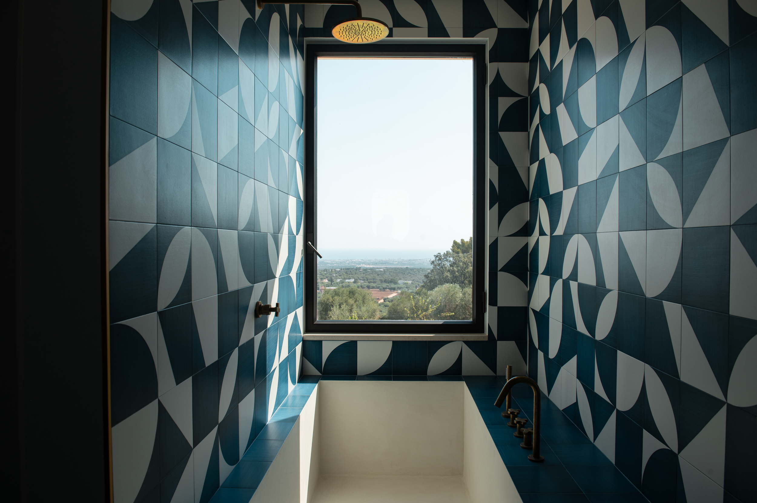 Bathroom with geometric blue and white tiled walls, a bathtub, a window with a scenic view, a rain shower ceiling fixture, and fixtures for a shower and faucet.